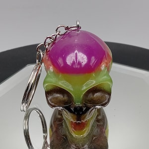 May include: A keychain featuring a resin skull figure with a smooth, glossy finish. The skull displays a gradient color scheme, transitioning from a vibrant magenta at the top to a translucent lime green in the middle and a dark brown at the base.