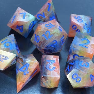 Pode incluir: Um conjunto de dados poliédricos coloridos com numeração azul. Os dados têm uma aparência marmorizada com tons de laranja, amarelo, azul e verde. O conjunto inclui várias formas, como D4, D6, D8, D10, D12 e D20.