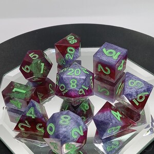 Könnte beinhalten: Ein Set aus polyedrischen Würfeln mit einer wirbelnden Mischung aus violettem und magentafarbenem Harz mit Glitzerakzenten. Jeder Würfel hat hellgrün gravierte Zahlen. Das Set enthält einen W20, W12, W10, W8, W6 und W4 auf einer reflektierenden Oberfläche.