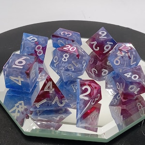 Op de afbeelding: Een verzameling polyhedrale dobbelstenen in verschillende vormen, waaronder D4, D6, D8, D10, D12 en D20. De dobbelstenen zijn transparant met een blauw-rood kleurenschema en zilveren cijfers. De dobbelstenen worden gepresenteerd op een spiegeloppervlak.