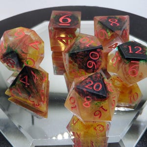 Pode incluir: Um conjunto de sete dados poliédricos para jogos de tabuleiro. Os dados são transparentes com um design de redemoinho amarelo e laranja, números vermelhos e detalhes em preto. Os dados são exibidos em uma superfície espelhada.