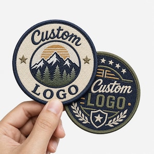Peut inclure: Deux écussons brodés circulaires avec des bordures bleu foncé. Le premier écusson présente un paysage de montagne avec un soleil, des arbres et le texte Custom LOGO. Le second affiche un bouclier, des étoiles, des lauriers et le texte Custom LOGO.