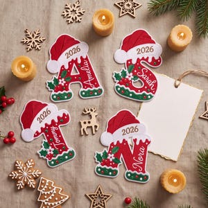 Puede incluir: Cuatro adornos con forma de letras A, S, T y N en rojo y blanco, cada uno con un gorro de Papá Noel y el año 2026. Las letras están decoradas con hojas de acebo verdes, bayas rojas y los nombres Amelia, Sarah, Tommy y Nora en letra blanca.