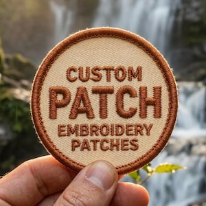 Könnte beinhalten: Ein kreisförmiger bestickter Aufnäher mit einer cremefarbenen Stoffbasis und einem dunkelbraun gestickten Rand. Der Text CUSTOM PATCH EMBROIDERY PATCHES ist in dunkelbraunem Garn mittig in einer fetten serifenlosen Schrift aufgestickt.