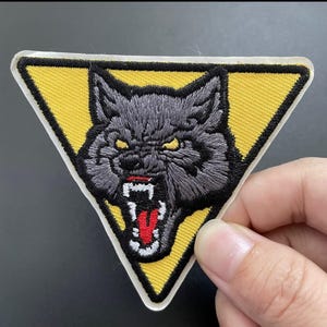 May include: A triangular embroidered patch featuring a snarling grey wolf head with yellow eyes and a red tongue. The wolf is centered on a bright yellow background with a thick black border. The design is stitched with detailed thread work.