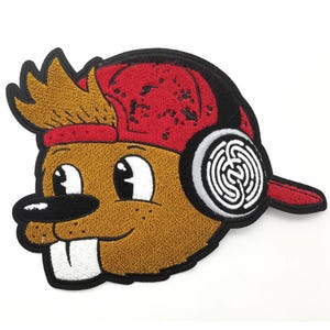 Könnte beinhalten: Ein bestickter Aufnäher mit einem Cartoon-Biberkopf mit braunem Fell, einer roten Baseballkappe, die verkehrt herum getragen wird, und weißen Kopfhörern mit einem schwarzen Labyrinth-Muster auf den Ohrmuscheln, großen Augen und zwei Zähnen.