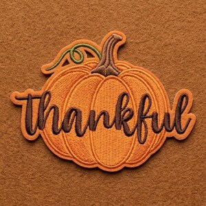 May include: An embroidered patch shaped like an orange pumpkin with a brown stem and a green vine. The word Thankful is stitched across the centre of the pumpkin in dark brown cursive script with a light brown outline on the pumpkin surface.