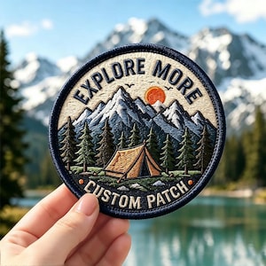 May include: A circular embroidered patch featuring a mountain landscape with a tent, pine trees, and an orange sun. The text EXPLORE MORE is arched at the top, and CUSTOM PATCH is arched at the bottom, all bordered by a dark blue stitched edge.