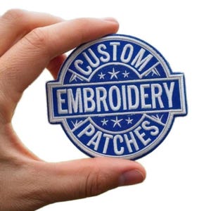 Op de afbeelding: Een ronde blauw-witte geborduurde patch met de tekst CUSTOM EMBROIDERY PATCHES in een cirkelvormige rand, voorzien van drie kleine sterren boven en onder de centrale tekst, alles ingesloten in een opvallend blauw-wit kader.