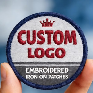 May include: A circular embroidered iron-on patch with a dark blue border. The top section is white with a red crown icon and the red embroidered text CUSTOM LOGO. The bottom section is dark gray with the white printed text EMBROIDERED IRON ON PATCHES.