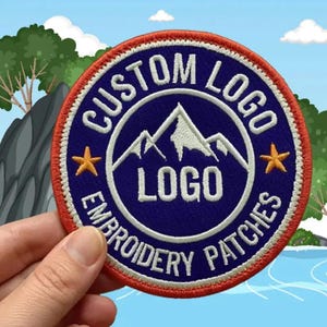 May include: A circular embroidered patch with a dark blue center and a bright orange border. The design features white mountain peaks, the text CUSTOM LOGO at the top, EMBROIDERY PATCHES at the bottom, and two small orange stars on the sides.
