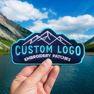 May include: A dark blue embroidered patch with a light blue border, featuring a white mountain outline graphic above the light blue text "CUSTOM LOGO" and the white text "EMBROIDERY PATCHES" in a curved, sans-serif font style.