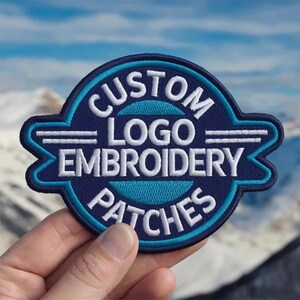 May include: A circular embroidered patch with a dark blue background and a bright blue border. The patch features the white text CUSTOM LOGO EMBROIDERY PATCHES arranged in a circular pattern with two horizontal white lines on each side.