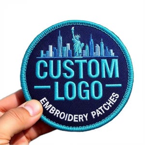 Könnte beinhalten: Ein runder gestickter Aufnäher mit dunkelblauem Hintergrund und hellblauem Rand. Das Design zeigt eine hellblaue Skyline-Silhouette inklusive der Freiheitsstatue, mit dem Text CUSTOM LOGO in Hellblau und EMBROIDERY PATCHES in Weiß.