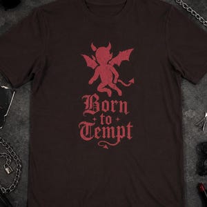 Puede incluir: Una camiseta de manga corta de color marrón oscuro con un gráfico rojo desgastado de un pequeño diablo alado sobre el texto "Born to Tempt" en una fuente de estilo gótico, con un pequeño detalle de cola de diablo al final.