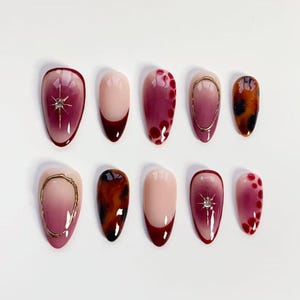 Könnte beinhalten: Ein Set aus zehn mandelförmigen Press-on-Nägeln mit verschiedenen Designs, darunter tiefrote Farbverläufe, nudefarbene Basen mit dunkelroten Spitzen, goldene Ringakzente, braune Schildpattmuster und kleine silberne Sternanhänger.