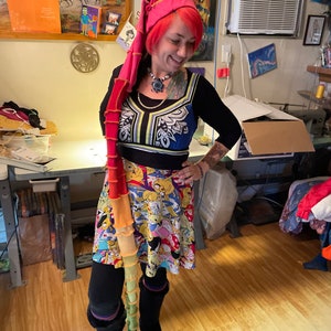 May include: A woman wearing a pink hat with a long, colorful, crocheted scarf. She is wearing a black and white patterned dress with a colorful cartoon print skirt. She is also wearing black boots.