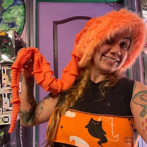 May include: A woman wearing a bright orange fuzzy hat and a black and orange Halloween themed shirt with a black cat and ghost graphic. She is holding a long, bright orange crocheted scarf.