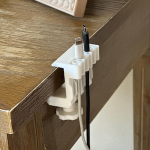 May include: A white plastic cable organizer with a clamp design, holding three charging cables. The organizer is attached to the edge of a wooden surface. One cable is white, one is black, and one has a lightning connector.