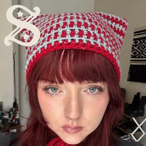May include: A red and light gray crochet kitty bonnet with pointed ears and long chin ties. The text on the image reads: kitty bonnet a crochet pattern by starlet studios. The bonnet features a checkered pattern and a solid red border.