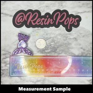 May include: A purple and iridescent glitter mermaid tail resin charm with a silver coin for size comparison. The charm is approximately 1 inch in length. The ruler in the background reads "Resin Pops" and is marked in inches.