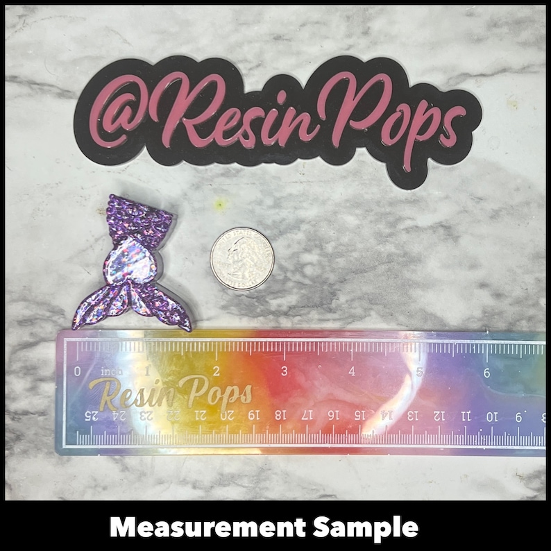 May include: A purple and iridescent glitter mermaid tail resin charm with a silver coin for size comparison. The charm is approximately 1 inch in length. The ruler in the background reads "Resin Pops" and is marked in inches.