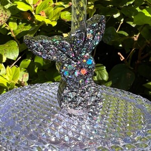 May include: A black glitter mermaid tail bottle stopper with iridescent glitter accents. The tail is sitting on a clear, textured surface.
