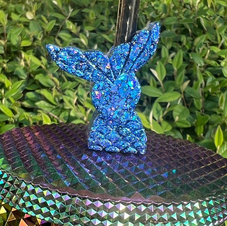 May include: A blue glitter bunny rabbit figurine with a sparkly finish. The figurine is sitting on a iridescent surface.