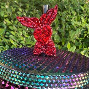 May include: Red glitter bunny shaped bottle stopper. The stopper is on a iridescent purple, green, and blue background.