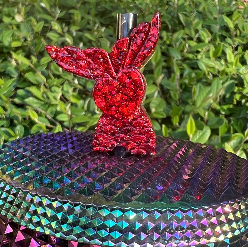 May include: Red glitter bunny shaped bottle stopper. The stopper is on a iridescent purple, green, and blue background.