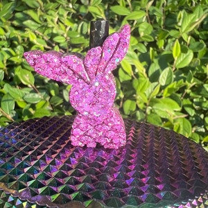 May include: Pink glitter bunny shaped bottle stopper. The stopper is sitting on a iridescent purple and green surface.