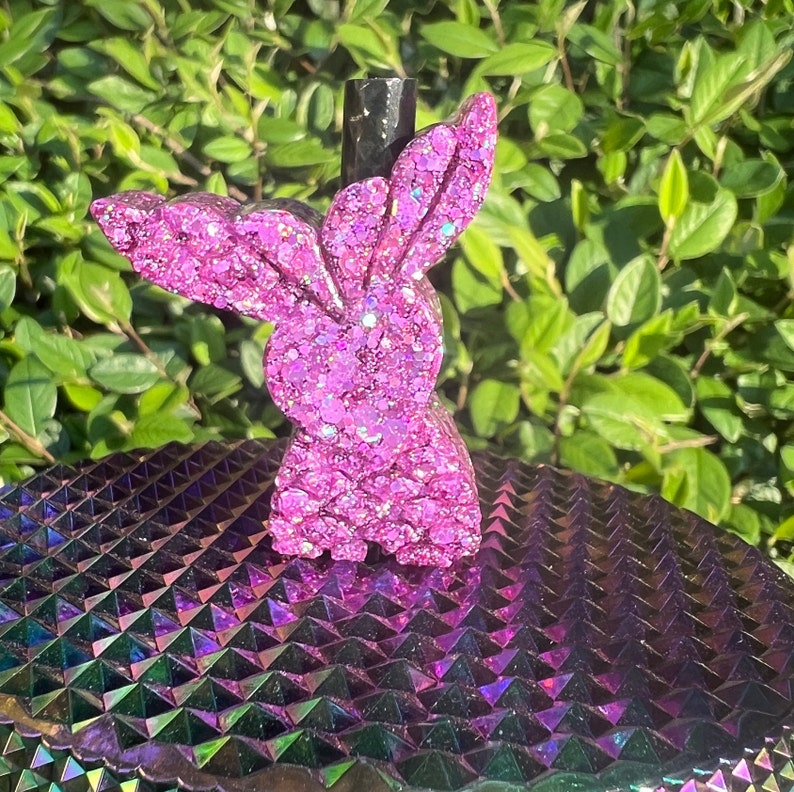 May include: Pink glitter bunny shaped bottle stopper. The stopper is sitting on a iridescent purple and green surface.