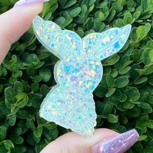 May include: A white iridescent glitter resin bunny charm. The bunny is facing to the right and has a large head and small body. The charm is made of clear resin with iridescent glitter embedded in it.