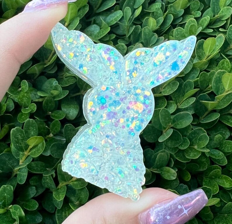 May include: A white iridescent glitter resin bunny charm. The bunny is facing to the right and has a large head and small body. The charm is made of clear resin with iridescent glitter embedded in it.