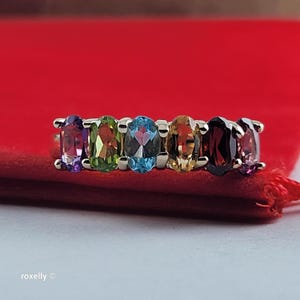 May include: A silver-toned ring featuring six oval-cut gemstones in a row, including purple, green, blue, yellow, dark red, and pink stones set in a metal band. The text "roxelly ©" is visible in the bottom left corner of the image.