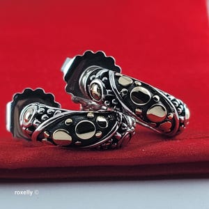 May include: A pair of hoop earrings featuring a two-tone design with polished silver-colored metal and smooth gold-colored circular accents set against a dark, oxidized background. The earrings have a textured, granulated surface detail.
