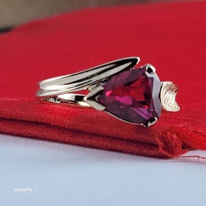 May include: A gold ring featuring a triangular-cut deep pink gemstone as the centerpiece. The band has a smooth, polished finish and curves toward the stone, which is flanked by a small, textured, leaf-shaped metal accent on one side.
