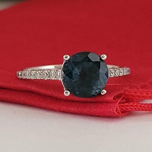 May include: A silver ring with a large, round, dark blue gemstone. The band is encrusted with small, clear stones. The ring is set against a red background.