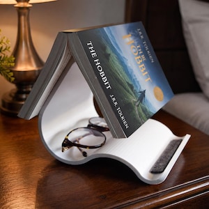 Op de afbeelding: Een witte, gebogen boekenstandaard van kunststof die een exemplaar van The Hobbit van J.R.R. Tolkien in een open, omgekeerde V-vorm houdt. Een leesbril met schildpadpatroon rust in de holle ruimte onder het boek met een landschapscover.