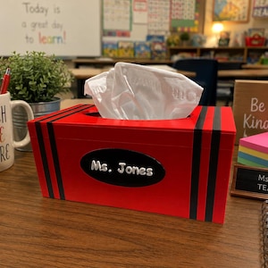 May include: A rectangular red tissue box cover designed to resemble a large crayon with black stripes at the ends and a black oval label on the front featuring the white text Ms. Jones. The box holds white tissues and sits on a wooden desk surface.