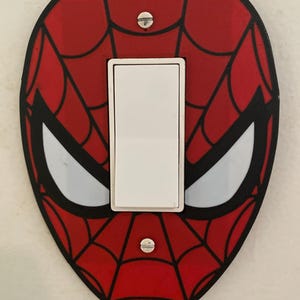 Puede incluir: Una placa de pared para interruptor de luz con diseño de Spider-Man en rojo y negro, con un patrón de telaraña y formas de ojos blancas. La cubierta ovalada tiene un corte central para un interruptor basculante blanco y dos tornillos.
