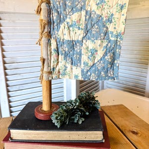 May include: A decorative flag made from a vintage quilt, featuring a blue and white floral pattern. The flag is attached to a wooden pole and sits atop a stack of old books. A sprig of faux greenery adds a touch of nature.