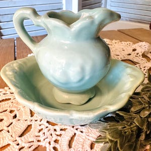 May include: A vintage, light blue ceramic pitcher and basin set. The pitcher has a curved handle and a scalloped rim. The basin has a matching scalloped edge. The set is displayed on a wooden surface with a lace doily.