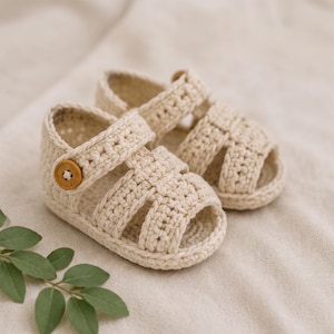 Può includere: Un paio di sandali all'uncinetto color bianco sporco con un design a cinturini intrecciati, punta aperta e un bottone rotondo in legno sul lato di ogni scarpa per la chiusura. Le calzature sono realizzate in filato spesso con aspetto materico.