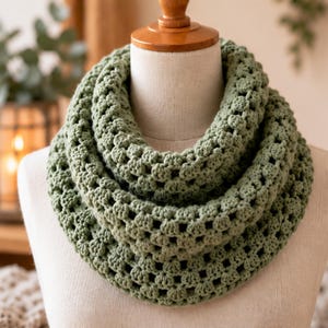 May include: A sage green infinity scarf featuring a textured crochet pattern with repeating clusters and open spaces. The scarf is draped in multiple loops around a beige mannequin neck, showcasing the consistent yarn work and color.