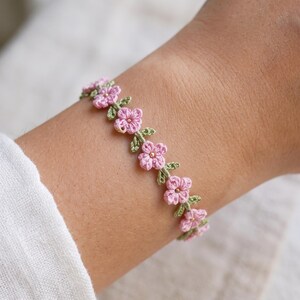 Peut inclure: Un bracelet délicat composé d'une série de petites fleurs crochetées rose clair avec des feuilles crochetées vertes. Chaque fleur est centrée par une minuscule perle dorée métallique, créant un motif de vigne florale continu autour du poignet.