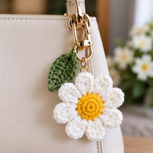 Könnte beinhalten: Ein gehäkelter Gänseblümchen-Taschenanhänger mit weißen Blütenblättern und einem gelben Zentrum, ergänzt durch ein einzelnes grünes Häkelblatt. Der Blumenanhänger ist an einem goldfarbenen Karabinerhaken befestigt, der an einer cremefarbenen Handtasche hängt.