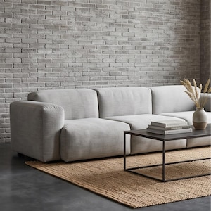 May include: A light gray modular sofa with a low-profile design and rounded armrests. A rectangular black metal coffee table sits in front, holding a stack of two books and a ceramic vase containing dried wheat stalks on a woven jute rug.