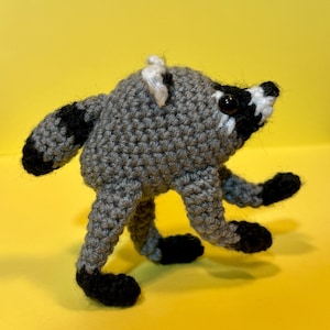 May include: A small, handmade crochet raccoon figurine crafted from grey, black, and white yarn. The plush toy features a rounded body, a striped tail, and black plastic eyes, with black yarn accents on the paws and facial markings.