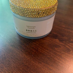 May include: A round jar with a gold, jewel-encrusted lid. The jar has a white label with the words "lightweight moisture" and a hydration level indicator. The jar is sitting on a dark brown wooden surface.